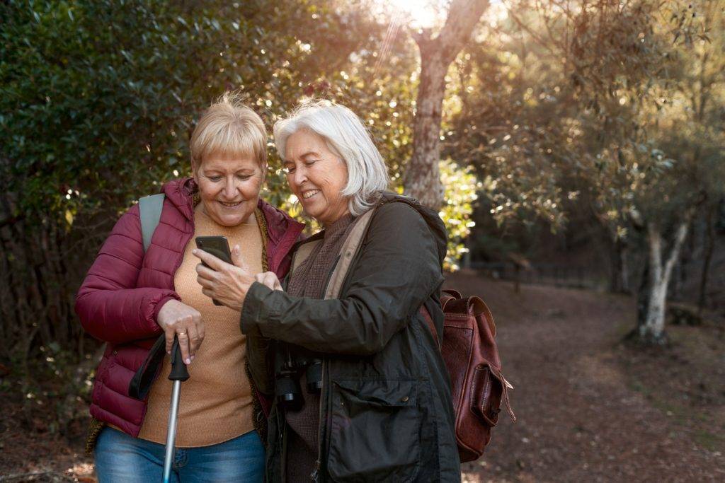 Aufnahme von zwei lächelnden Seniorinen bei dem Betrachten eines Bildes auf einem Handy. Sie sind auf einem Auflug in einem grünen und lichtem Wald.