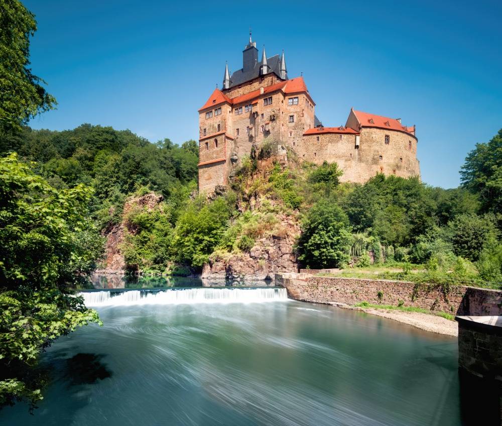 Panorama Blick auf einen tiefblauen Fluss mite einem kleinem Wasserfall, grüne Baume, Sträucher und Wiese und in Hintergrund eine hohe Burg mit rotem und schwarzem Dach.