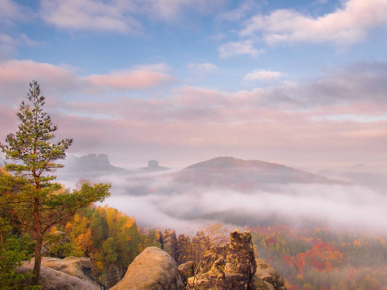Ein atemberaubender Blick auf eine vernebelte Berglandschaft mit bunten herbstlichen Bäumen. Im Vordergrund ine felsige Ebene mit vielen herbstlichen Laubbäumen.